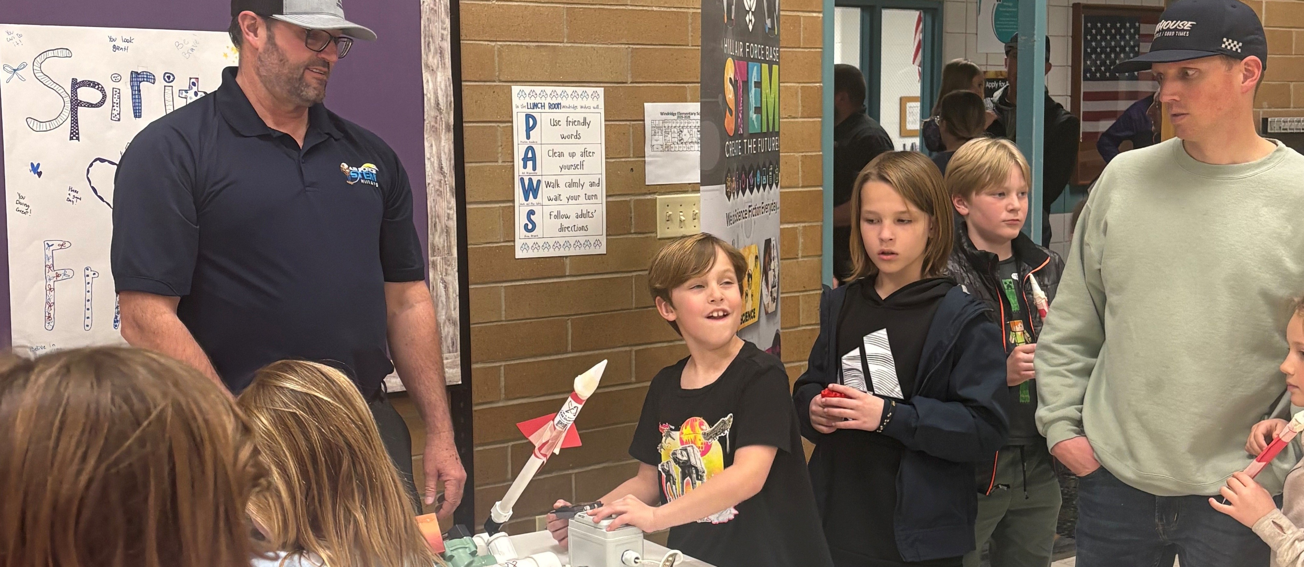 Boy shooting a paper rocket off a table device while kids and parents watch.