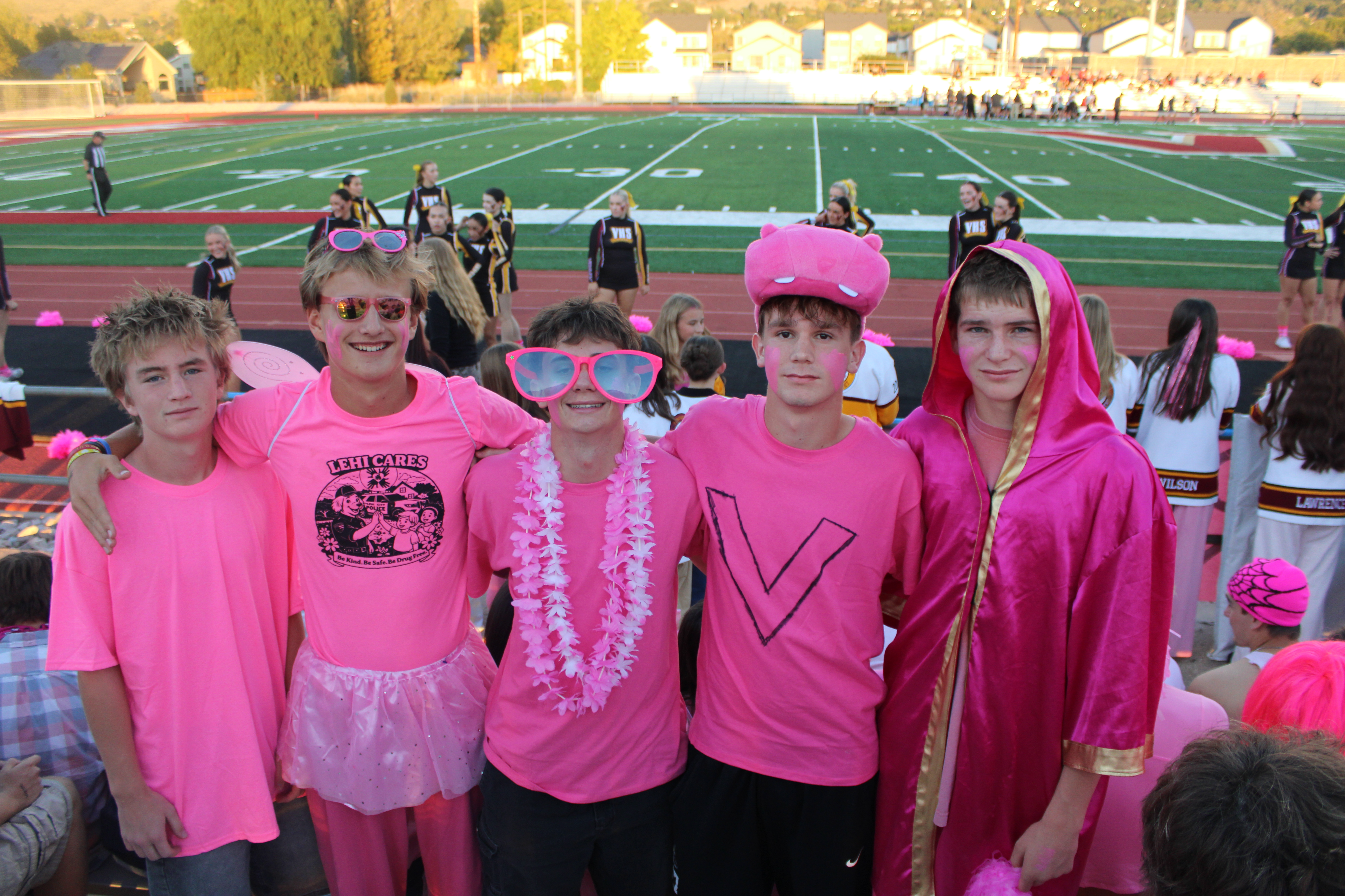 Five high school students pose together in bright pink outfits at a football game’s pink-out event. Their outfits include oversized pink sunglasses, a tutu, a flower lei, a plush pink hat, and a shiny pink robe. Cheerleaders and spectators gather behind them along the track, with the football field in the background.