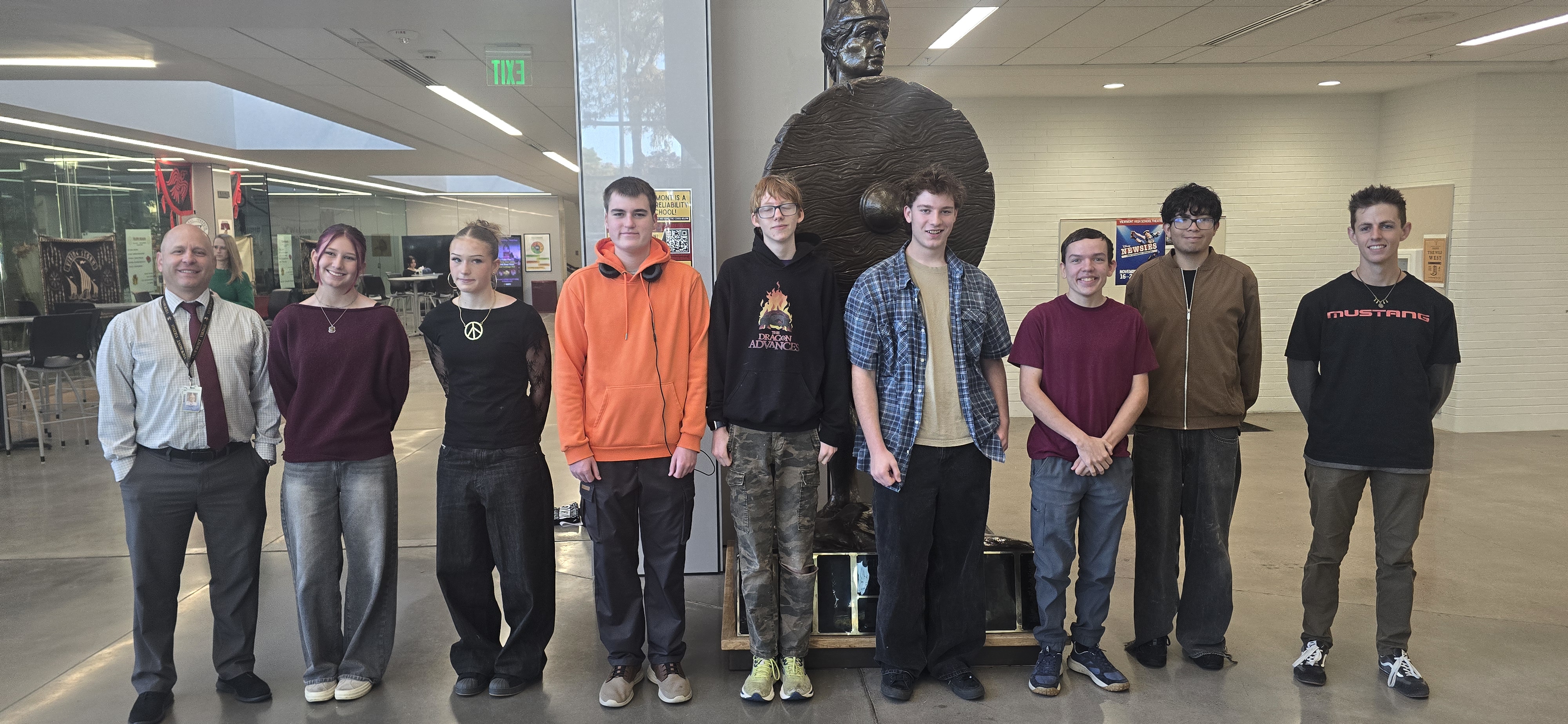 A group of nine people, including eight students and one adult, pose for a photo inside a university building. They stand in front of a large bronze statue, smiling at the camera in a spacious lobby with glass walls and overhead lighting.