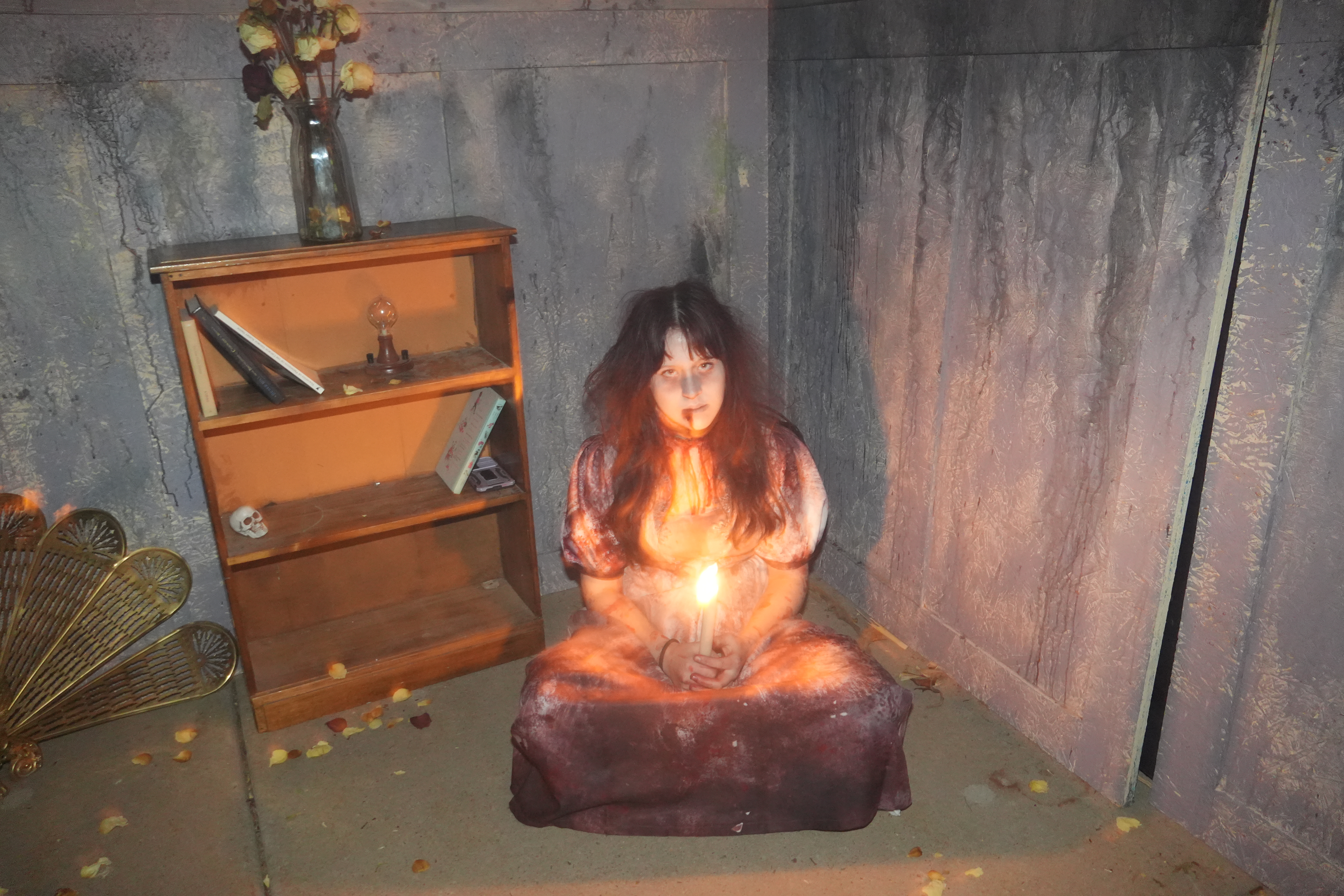 Person with long dark hair sits cross-legged on the floor of a dimly lit, gray room, holding a glowing candle. The scene includes a wooden bookshelf with books, a skull decoration, and a vase of dried roses. Rose petals are scattered on the floor, and the candlelight casts warm highlights against the dark, textured walls, creating a spooky, gothic atmosphere.