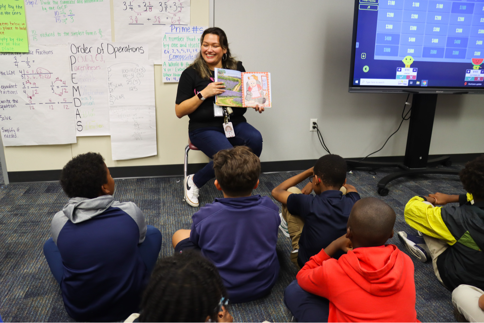 Woman sitting in front of students on a stool, holding a book. Monitor and whiteboard in the background.