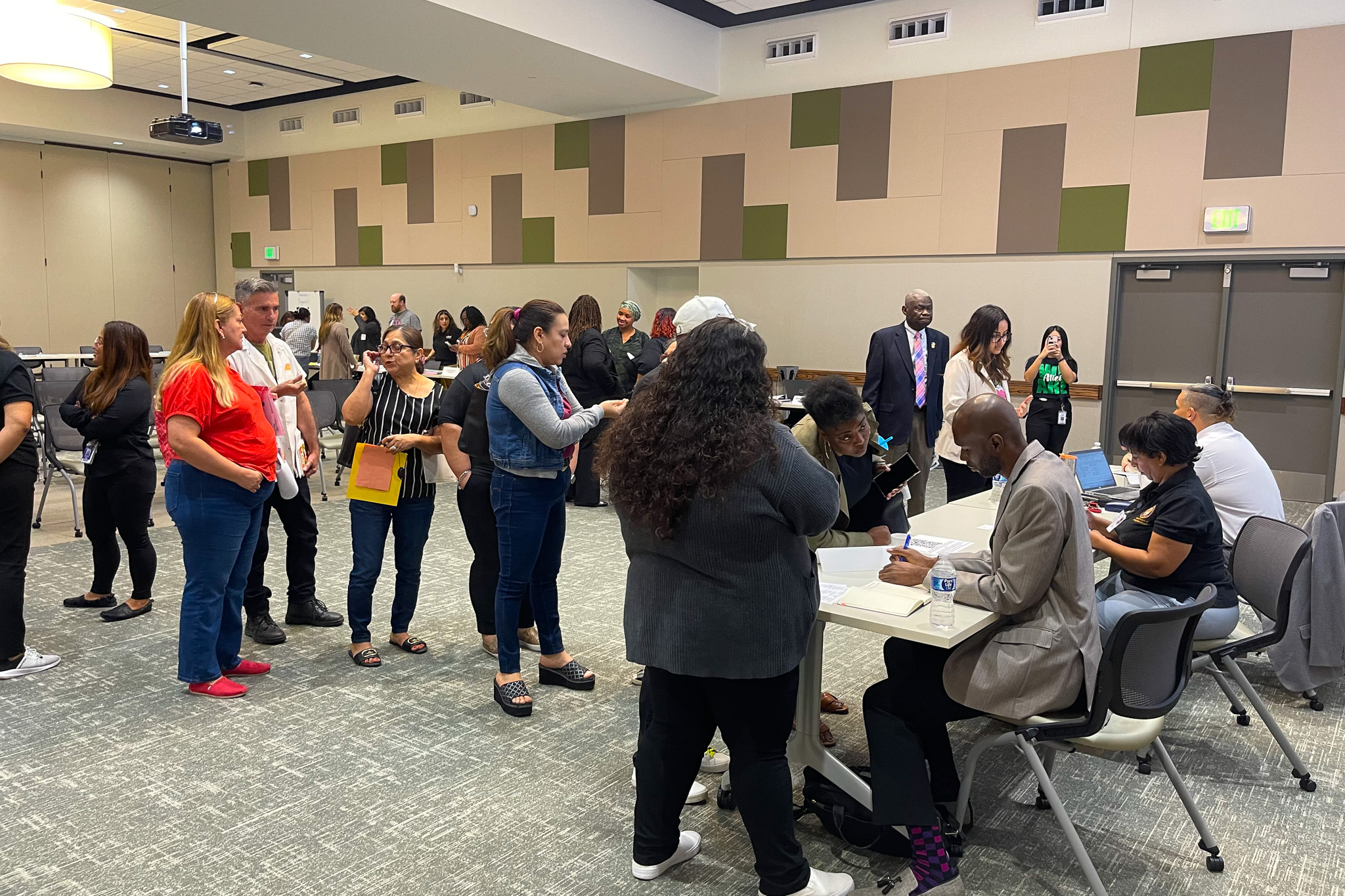 People stand in groups inside a room with tiled flooring, and some sit at tables with papers.