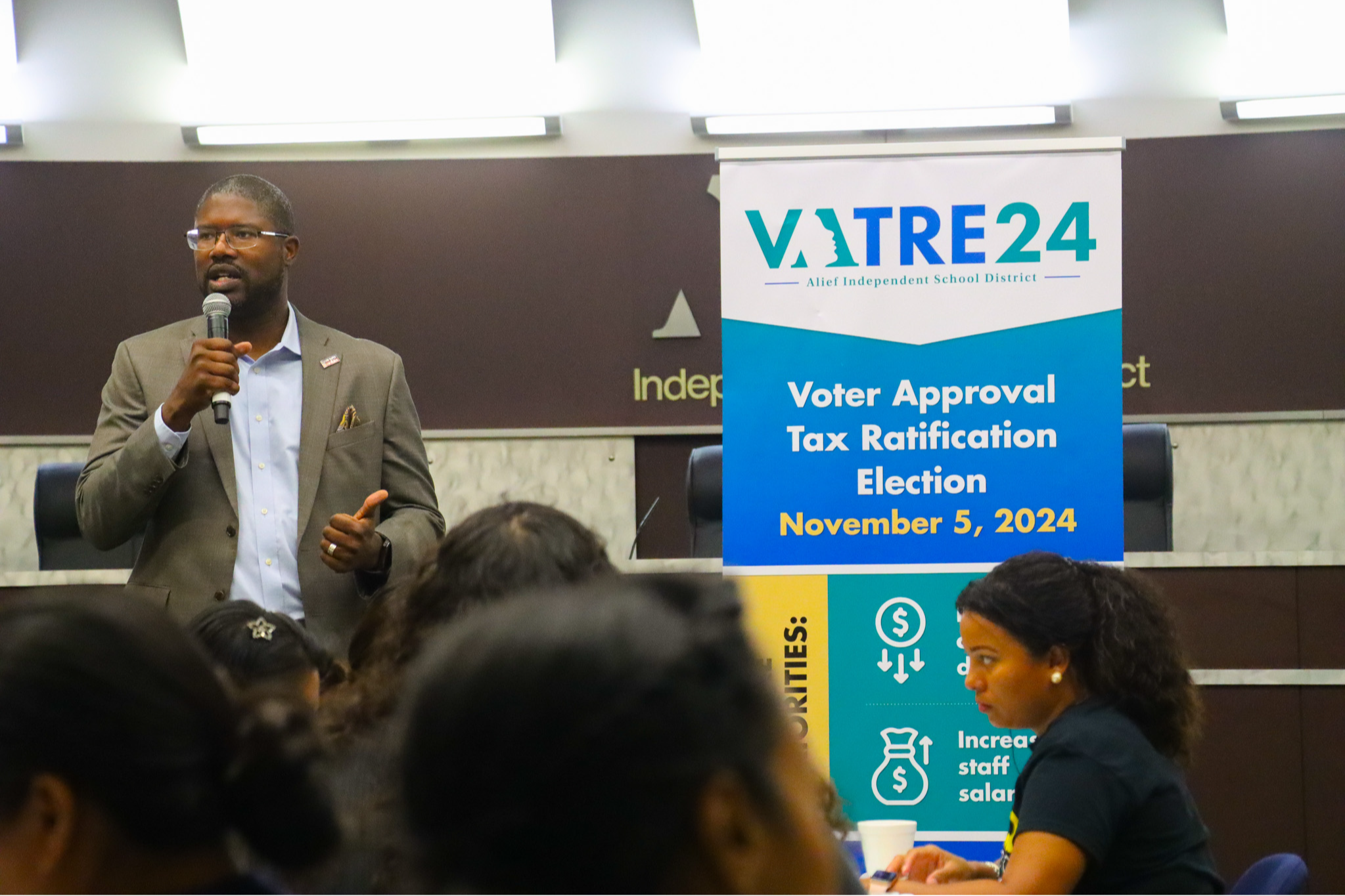 A man speaks into a microphone at an event. Banners are behind him, one titled "Voter Approval Tax Ratification Election November 5, 2024".