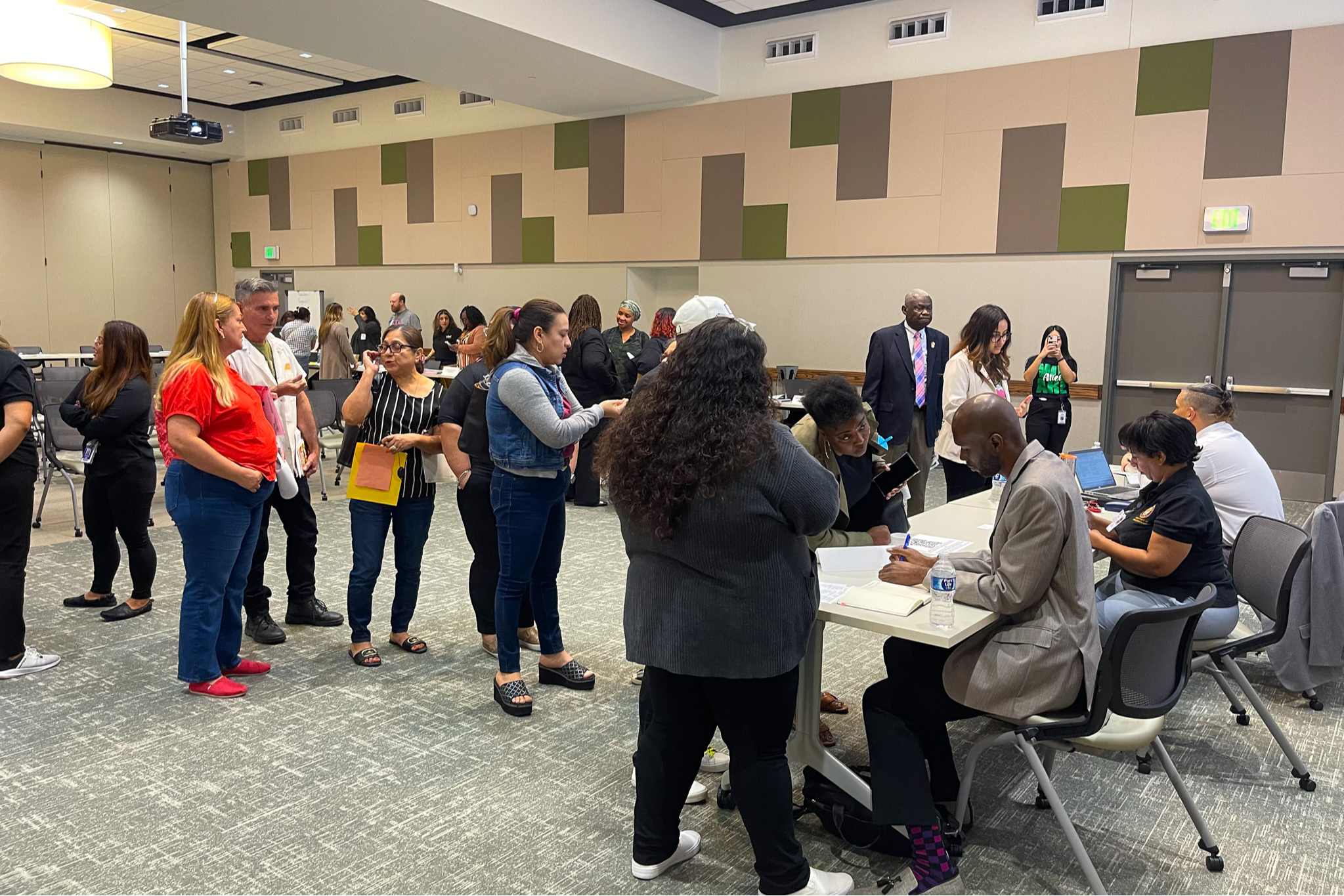 People stand in groups inside a room with tiled flooring, and some sit at tables with papers.