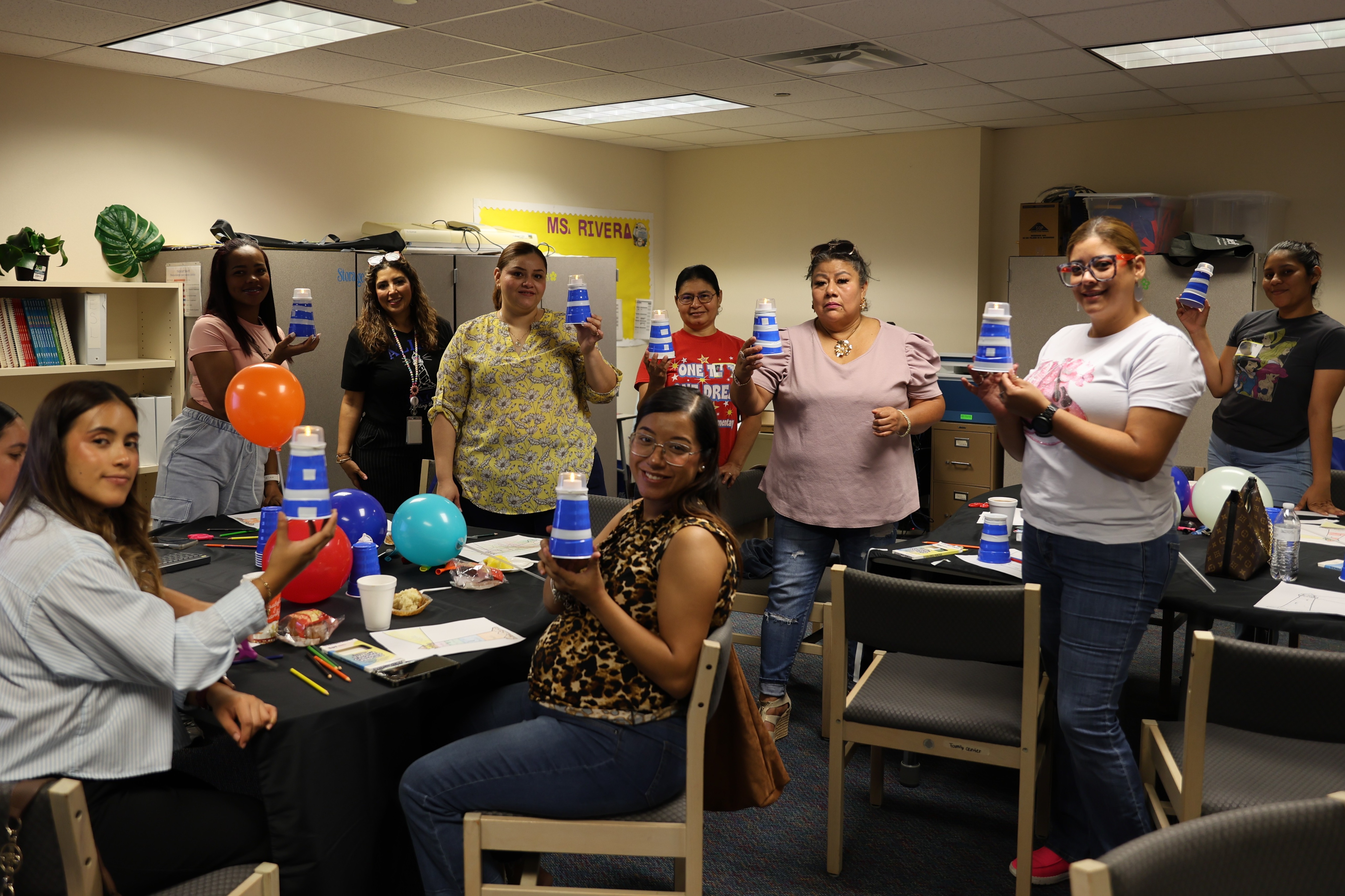 A group of people in a room with tables, chairs, and balloons. Some hold cups, and others hold cups with lids.