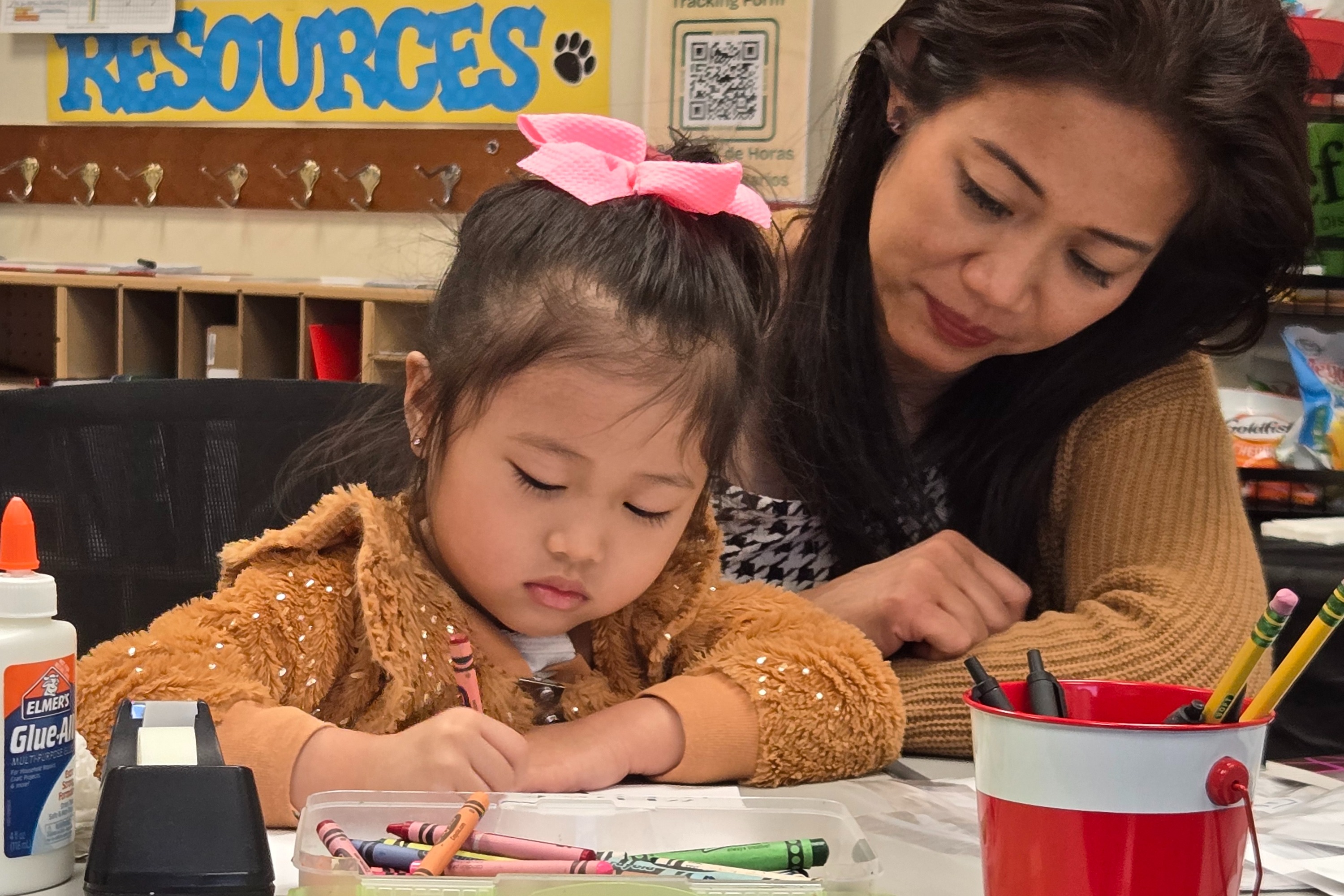 A young girl with a pink bow sits at a desk, drawing while a woman watches.