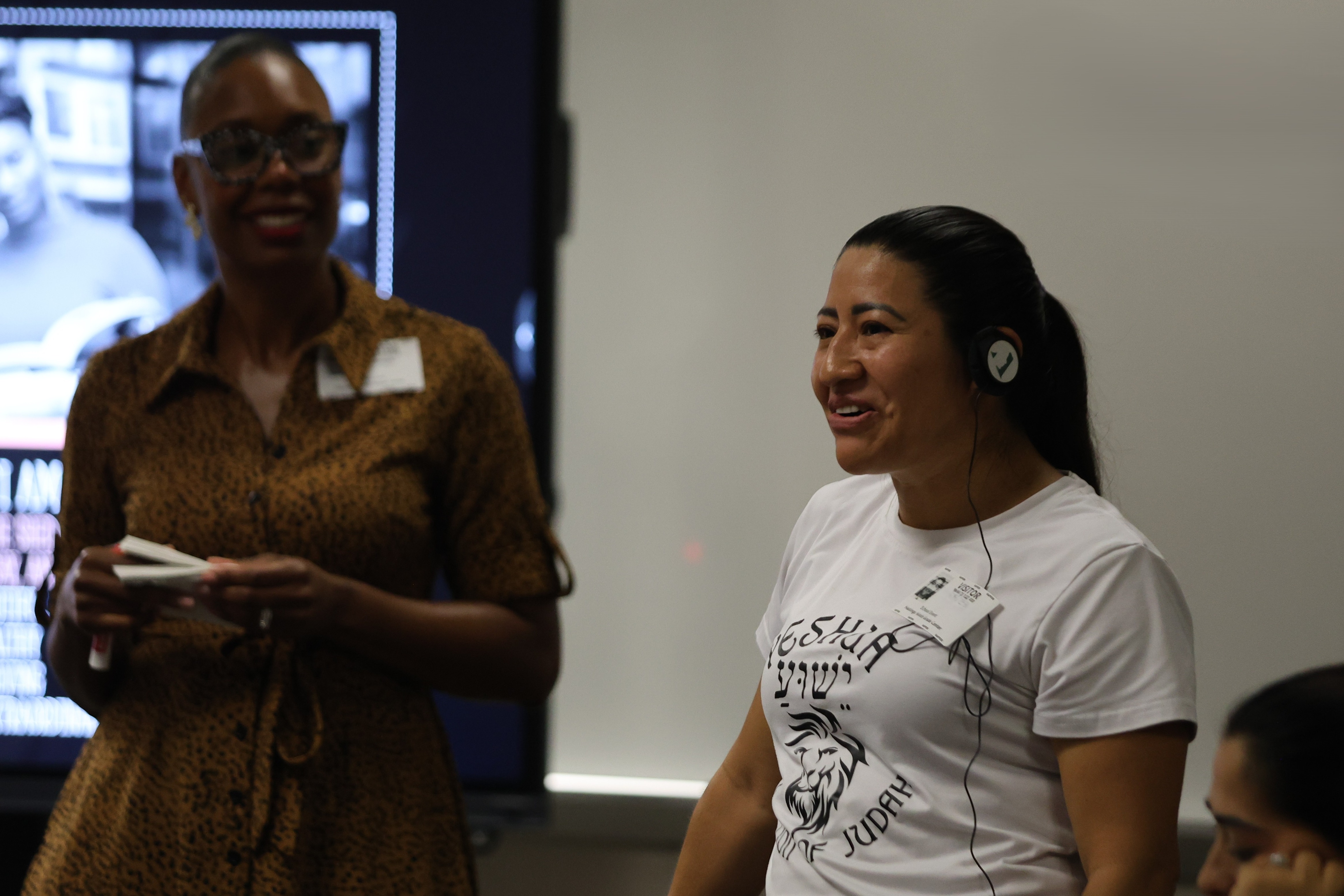 Two women interact in a classroom. One holds a remote control, while the other wears a white shirt. A screen displays a photo.