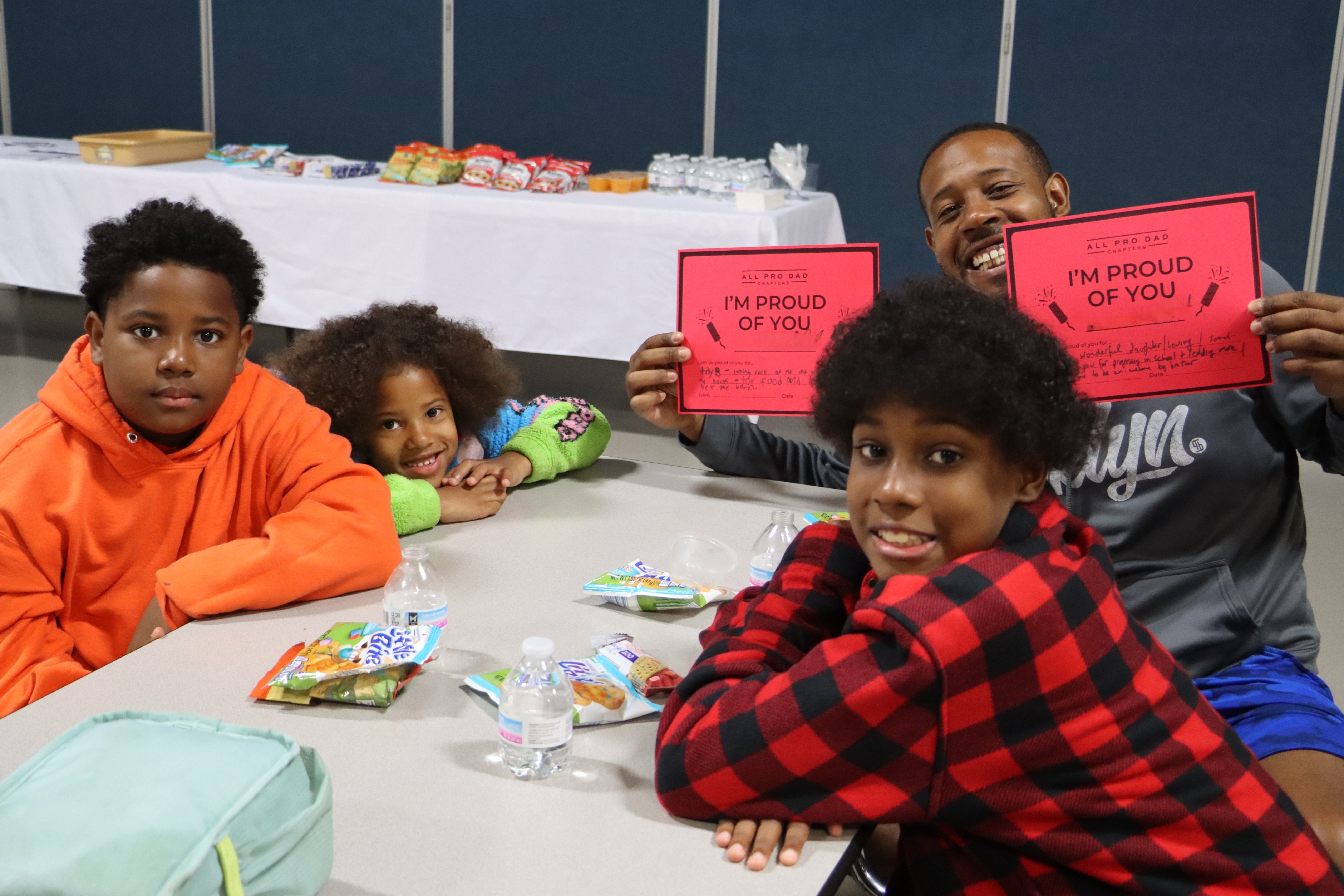 Four children and an adult sit around a table, smiling. The adult and one child hold up signs that say "I'm proud of you."