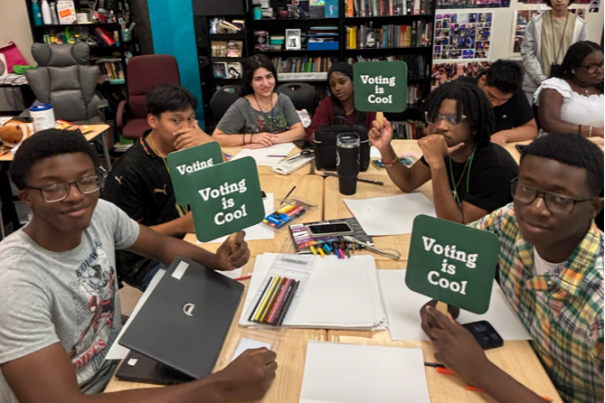 Group of students around a table in a classroom, holding signs that say "Voting is Cool."