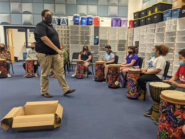 A man playing drums in a classroom surrounded by other people engaged in musical activities.
