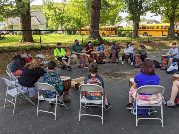 Several individuals seated in chairs around a drum, engaged in a discussion or performance.