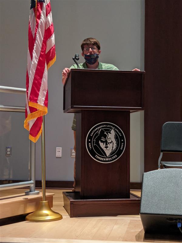 A man wearing a mask stands at a podium with an American flag, symbolizing a public address during uncertain times.