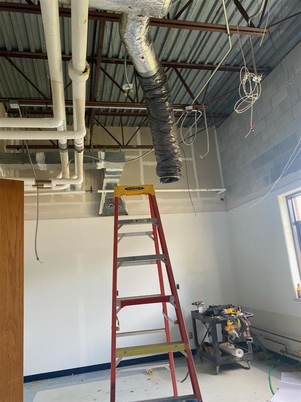 The image shows a ladder in the middle of a room with a ceiling fan. The ladder is a step ladder, which means it has flat steps that a person can stand on. The ladder is leaning against a wall, with the top of the ladder near the ceiling fan. The ceiling fan has four blades and is hanging from the ceiling by a metal pole.
