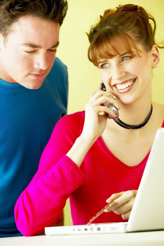 A man and woman having a conversation on a cell phone while gazing at a laptop screen.