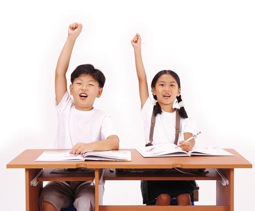 Two children sitting at a desk with their hands up, eagerly participating in a classroom activity.