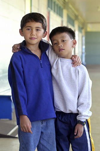 Two young boys standing in a hallway, wearing backpacks and smiling.