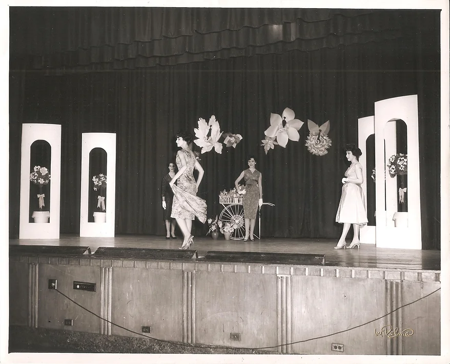 black and white 1950s photo of four women standing onstage at what appears to be a fashion show