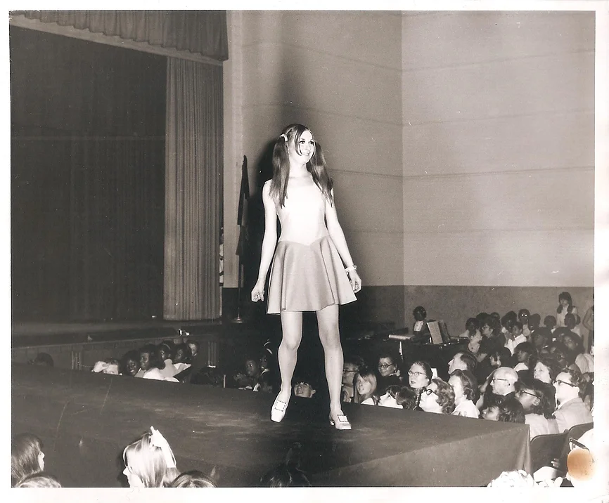 black and white 1960s photo of a model in pigtails and a thigh-length a-line dress on a runway