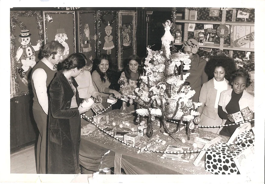 black and white 1970s photo of five friends candidly smiling around a mini christmas tree