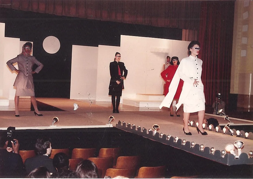 five female models posing on a runway in suit-dresses and futuristic sunglasses, in the 1980s