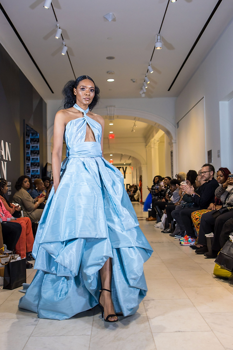 female model of color in a floor length blue gown walking down a runway of people