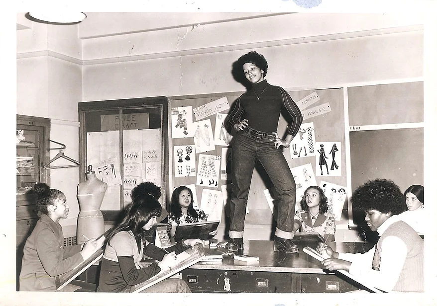 black and white 1070s photo of a live model of color posing with hands on hips on a table while students sit at her feet and draw her
