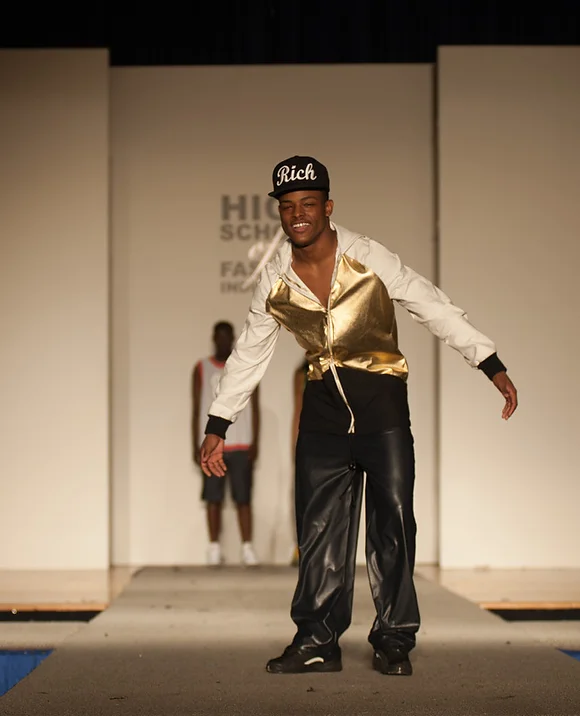 male model of color posing on a runway wearing a shiny gold zip up hoodie, baggy black leather pants, and a black hat that says rich