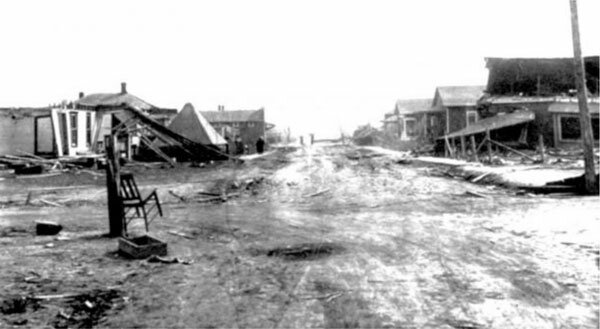 Black and white photograph showing a dilapidated town after a tornado, with debris scattered across the street. The contrast between the desolation in the foreground and the resilience of the standing structures in the background suggests the aftermath of a natural disaster.