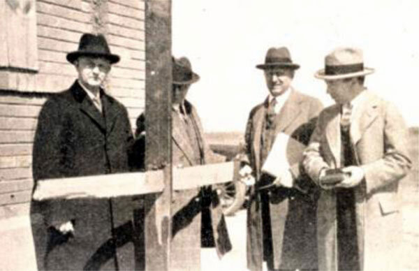 A group of four men, dressed formally in suits and ties, standing next to a wooden sign post at the entrance of what appears to be an event or gathering. 