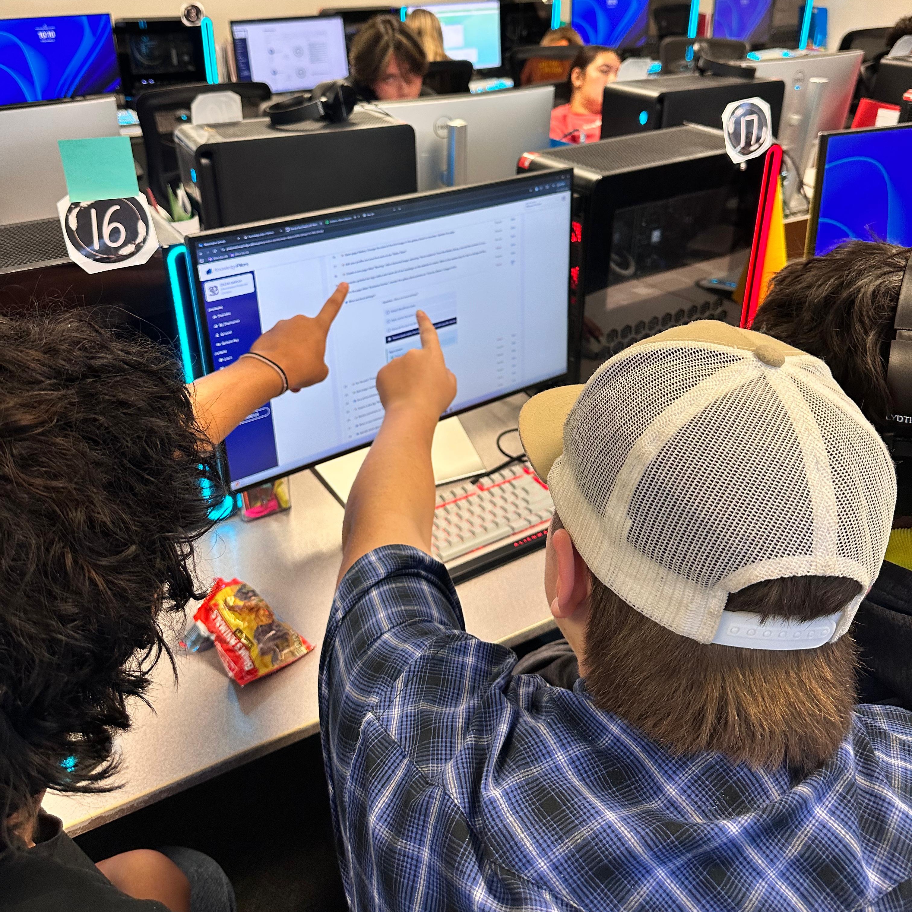 People using computers. One man points at a monitor screen. He wears a hat. A wall holds posters.