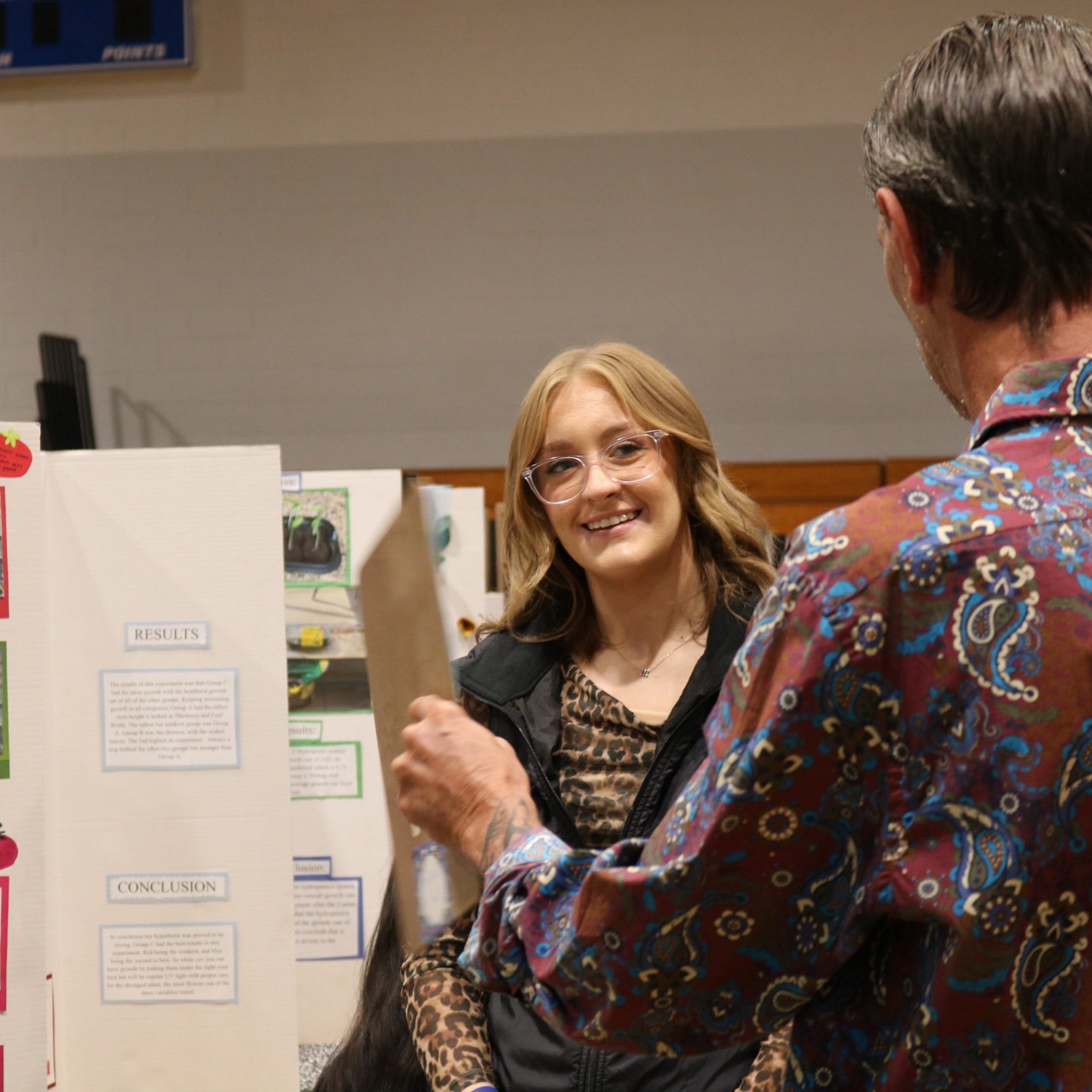 Two people standing, one holding paper, in front of a bulletin board with pictures and text.