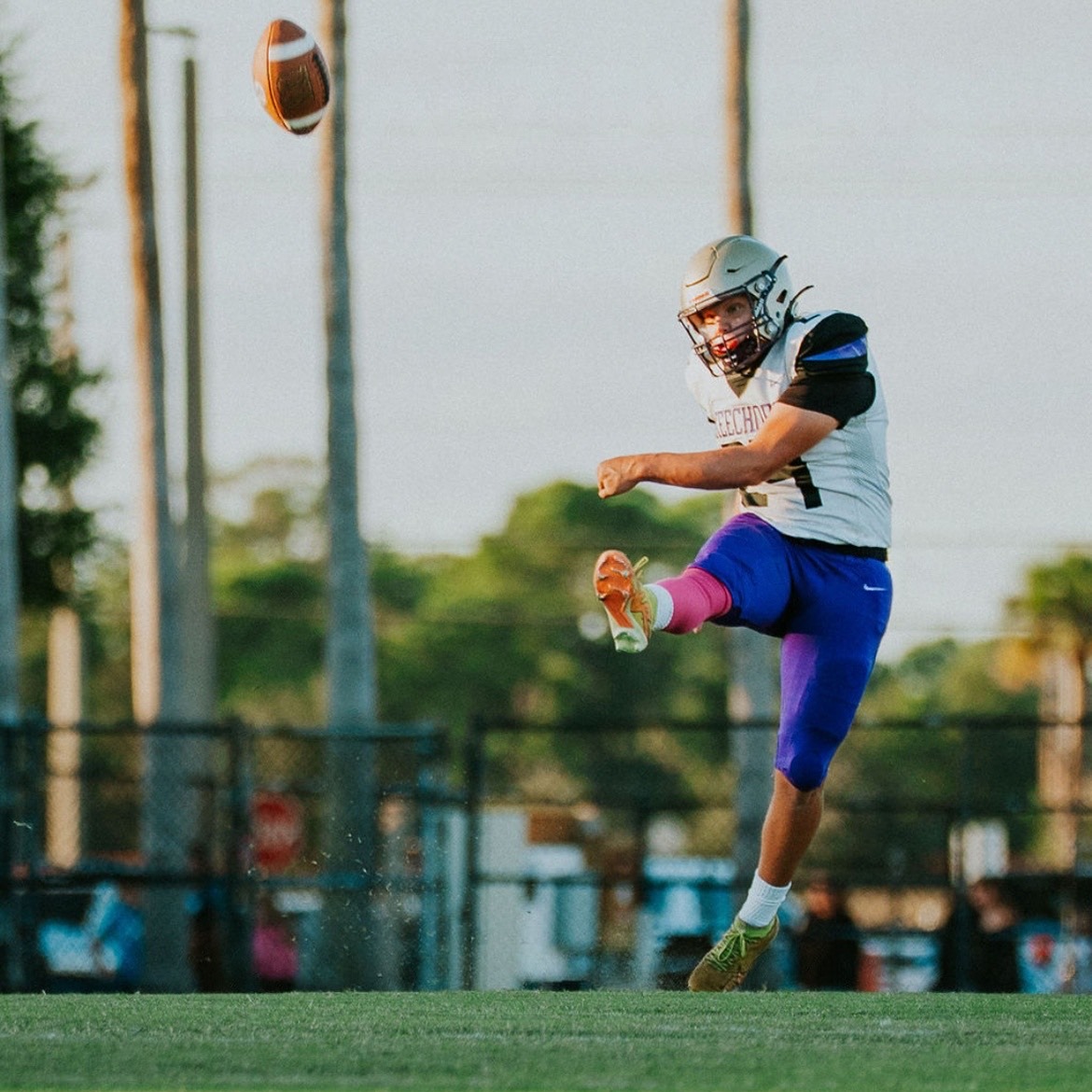 A football player in a white jersey with number 1, blue pants, and sneakers kicks a ball in midair.