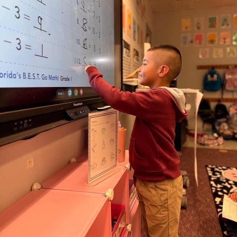 A young boy pointing at an interactive whiteboard displaying math problems. The classroom has a pink cabinet and posters.