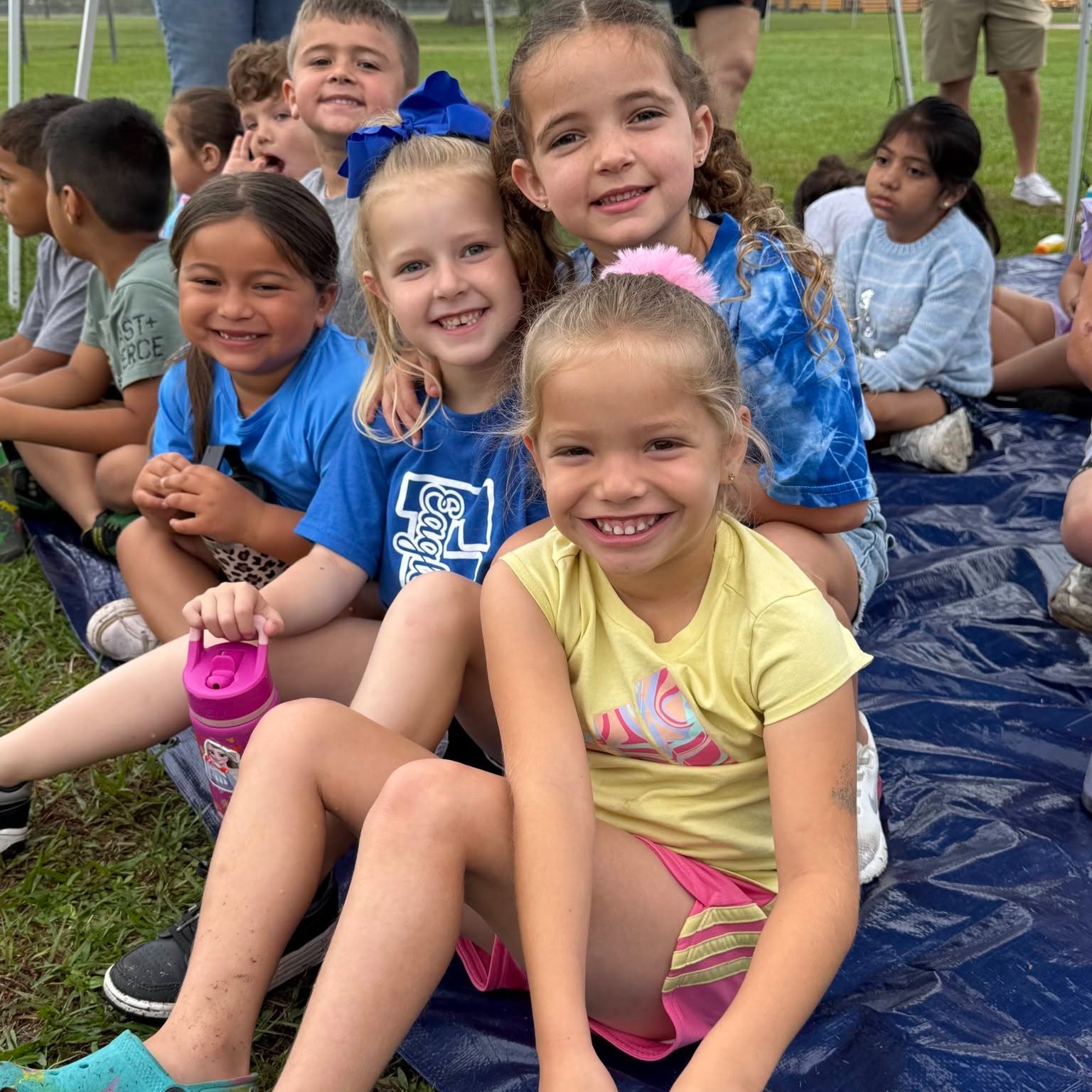 Children in colorful clothing sit on a blue tarp on grass, smiling and posing for a photo.