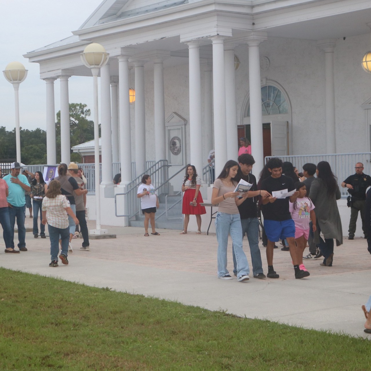 Multiple people stand in a line in front of a white building with columns, a red brick building in the background.