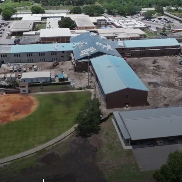 Aerial view of a construction site featuring multiple buildings, some with blue roofs. Green grass, a dirt field, and a body of water in the background.