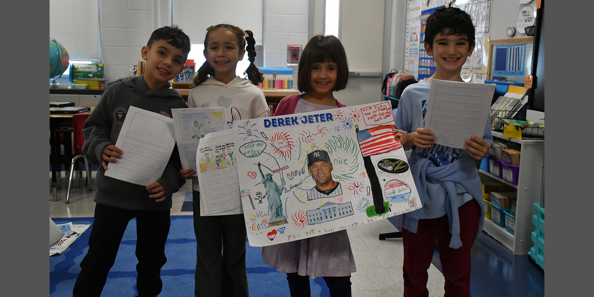 children stand in a classroom, holding projects and smiling. 