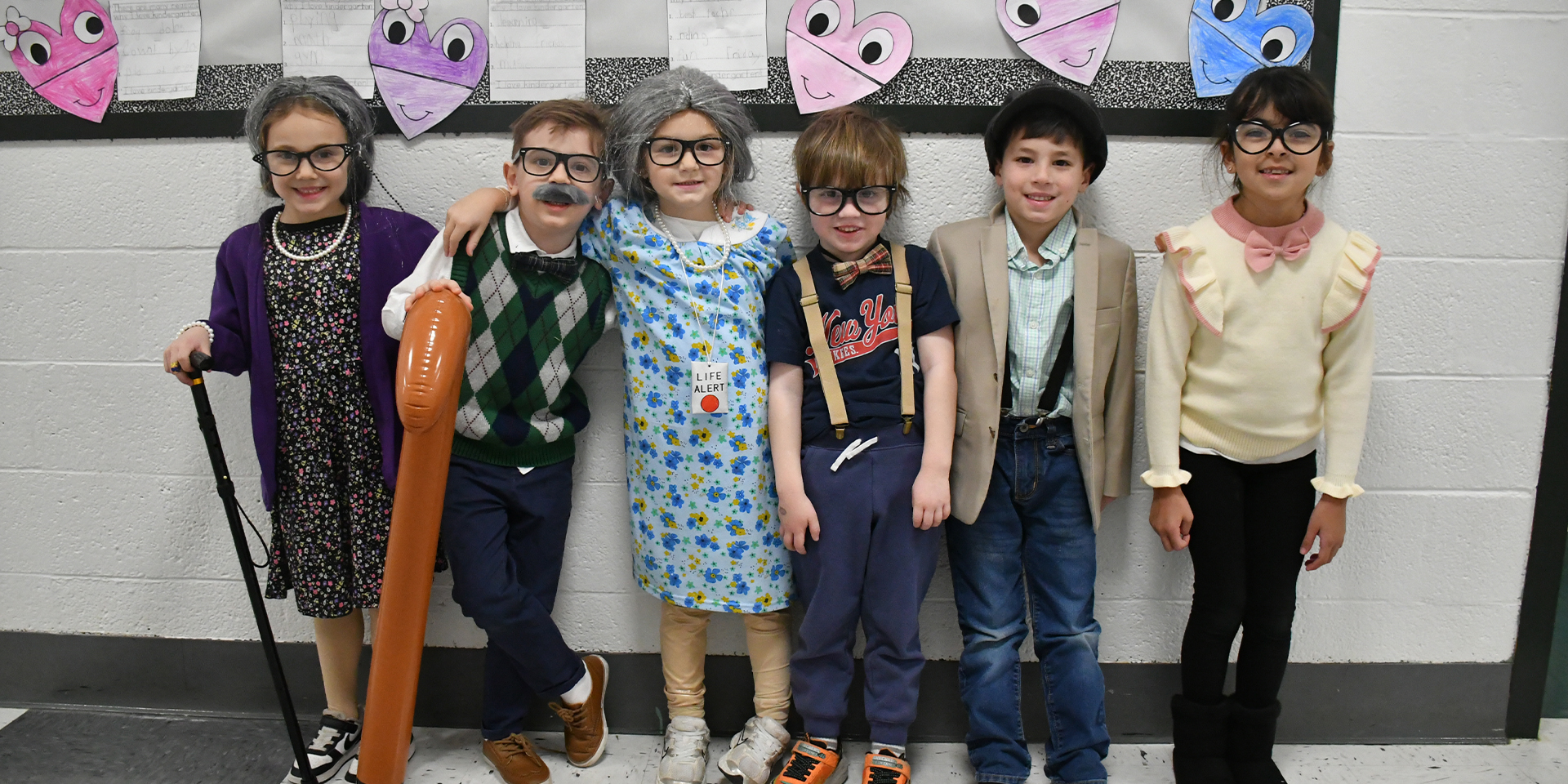 Six children are dressed as elderly people for a school event, wearing glasses, wigs, and costumes. They stand smiling against a wall decorated with heart-shaped drawings, creating a playful and fun atmosphere.