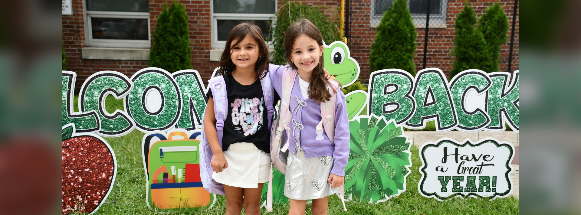 Two girls stand in front of a colorful “Welcome Back” sign with school-themed decorations