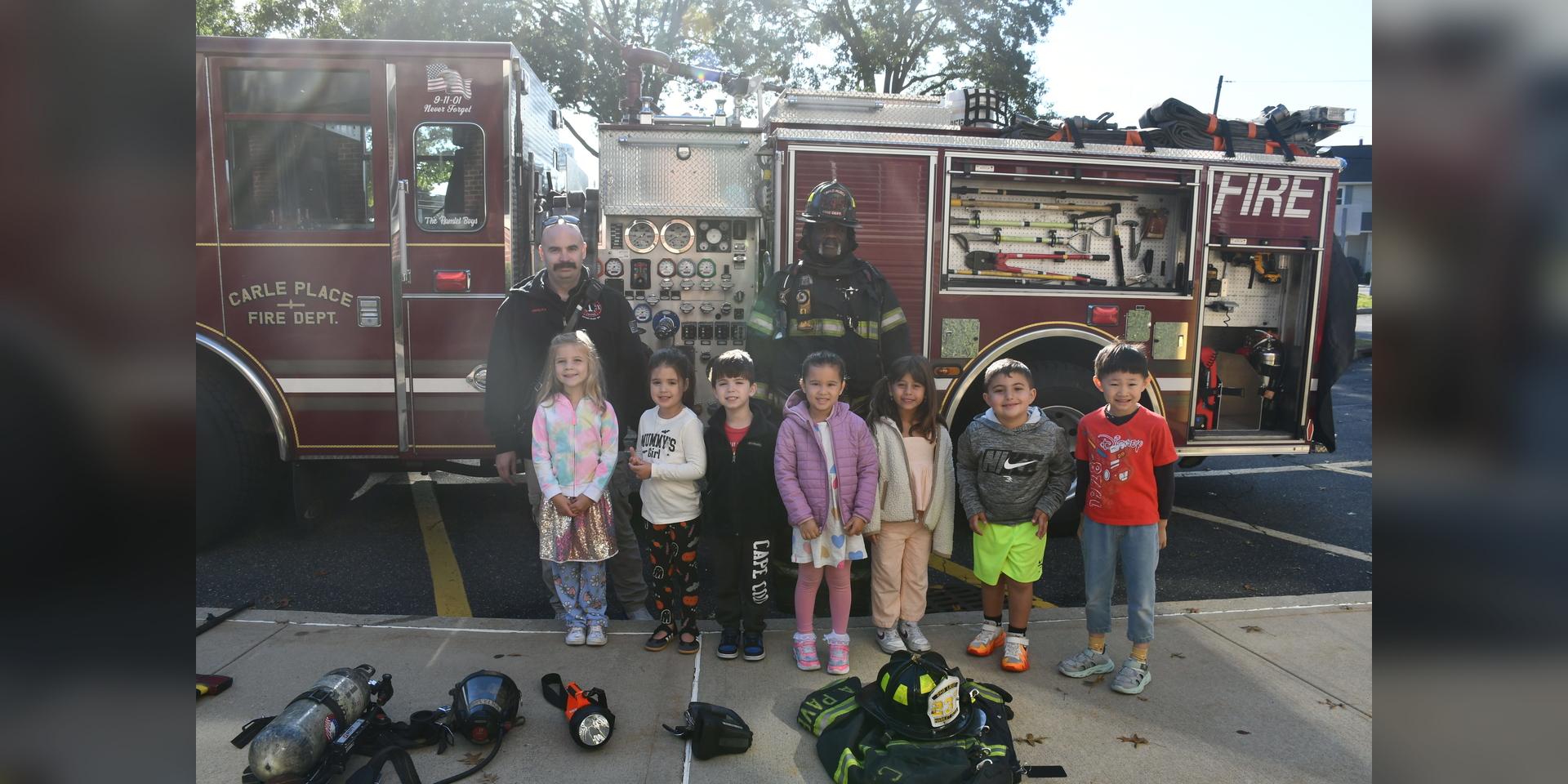 A group of children poses with a firefighter outside a trailer and parked vehicles.