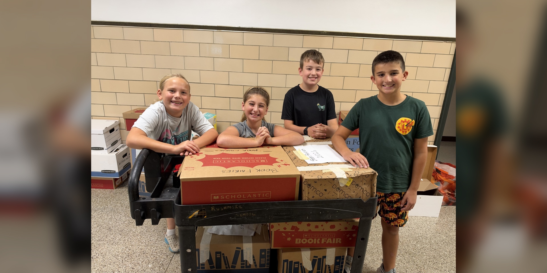 A group stands behind a cart loaded with boxes in a school hallway.