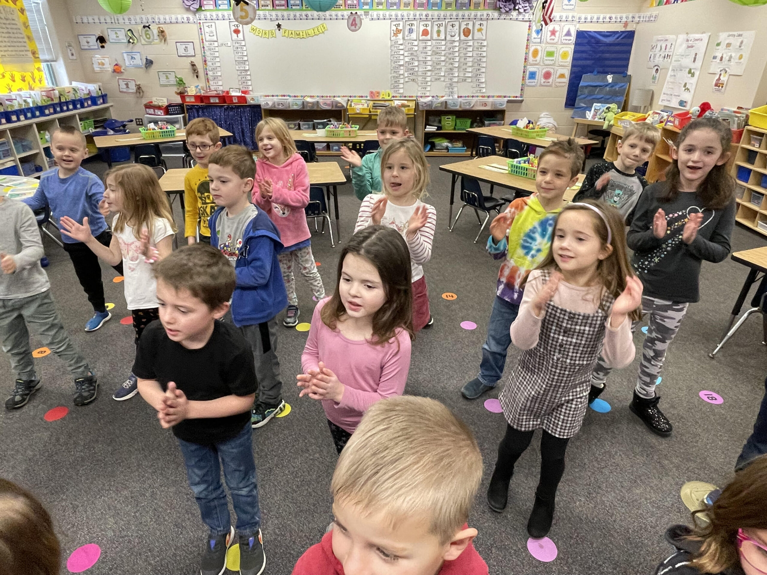 Children in a classroom attentively listening to their teacher as they stand in a circle.