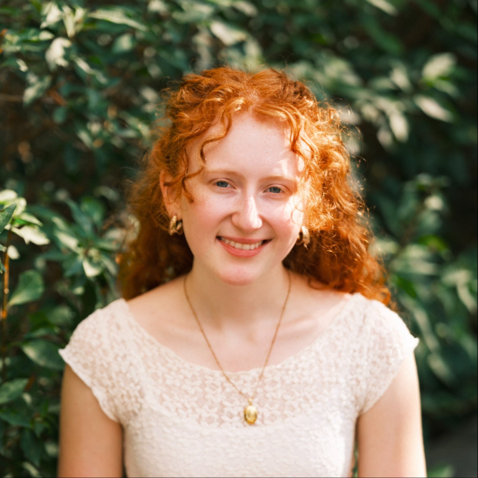 Zoe LeFeber - Person with red curly hair wearing a white blouse and gold necklace sits in front of greenery.