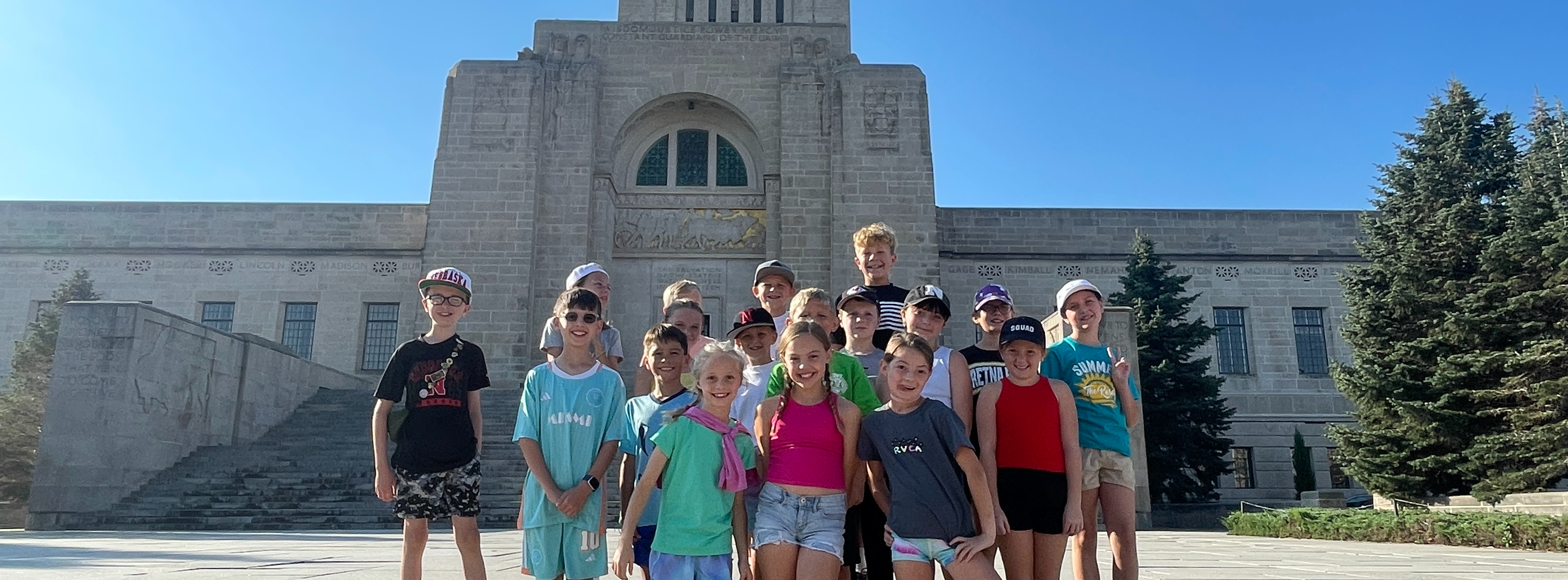 A group of smiling children, appearing to be between 8 and 12 years old, stand on the grand stone steps of a large, imposing government building under a clear blue sky. The building, possibly a capitol or courthouse, features classical architectural elements and a prominent central tower. The children are dressed in casual summer clothing, including t-shirts, shorts, and caps, and are arranged in a few rows, looking directly at the camera. Some children have their arms around each other or are making playful gestures. Green evergreen trees are visible on the right side of the frame, complementing the gray tones of the building and steps. The overall impression is one of a school trip or outing, capturing a moment of youthful energy and enthusiasm in front of a significant landmark.