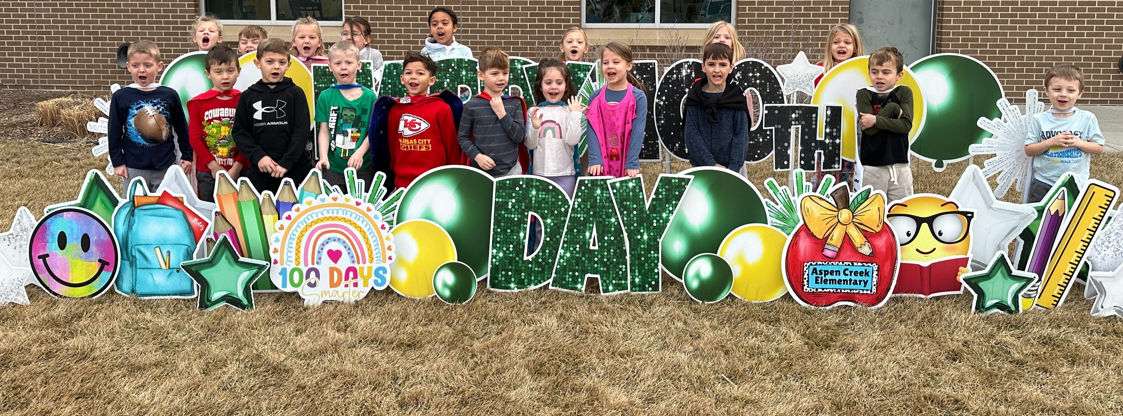 A large group of approximately twenty young elementary students is posed outdoors in front of a brick school building to celebrate their 100th day of school. The children are standing behind and around an elaborate, oversized yard sign display.