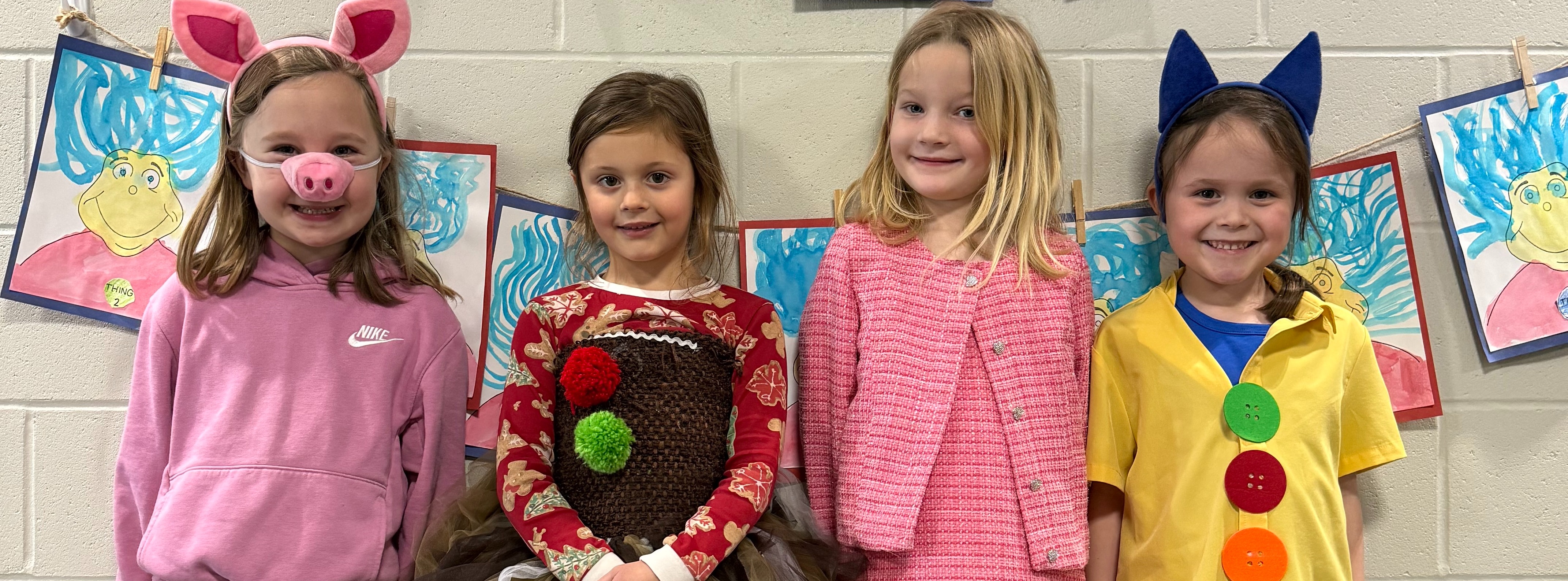 Four young girls stand in a row against a classroom wall, dressed in various costumes. From left to right: a girl in a pig nose and ears, a girl in a brown tutu with festive patterns, a girl in a pink dress and cardigan, and a girl in a yellow shirt with large colorful buttons and blue cat ears. Behind them, a string of hand-drawn "Thing 1 and Thing 2" characters is displayed.