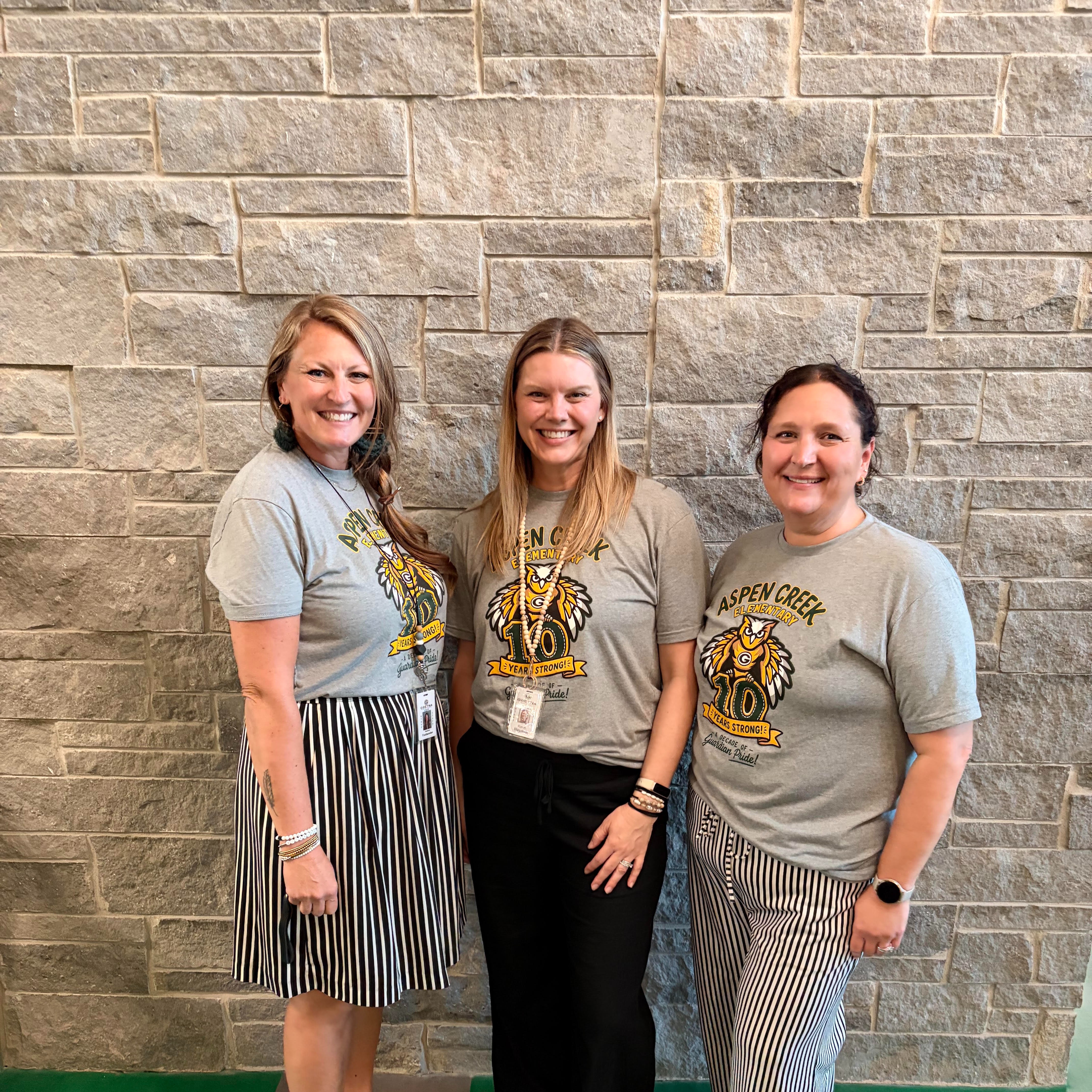 Three women smiling together in matching grey "Aspen Creek Elementary" t-shirts in front of a stone wall.