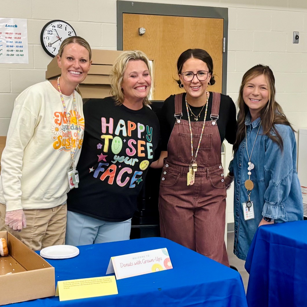 Four smiling women standing together behind a blue table with a sign reading "Donuts with Grown-Ups."