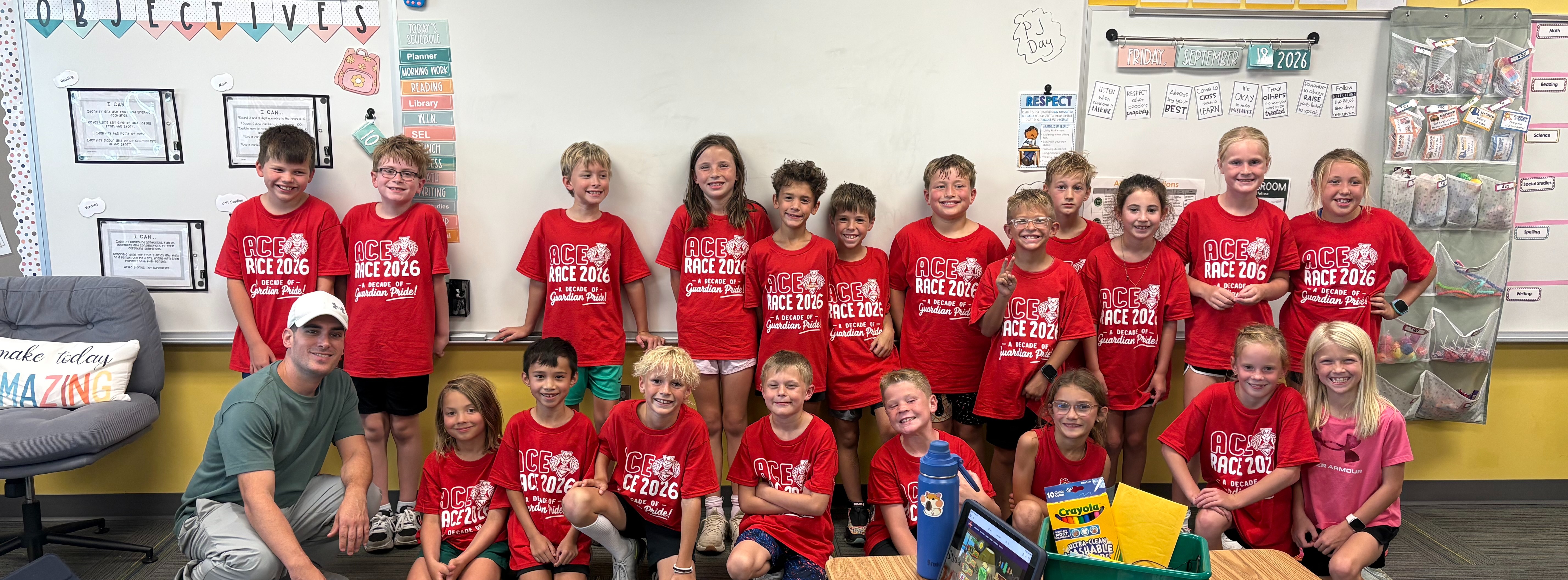 An adult male kneeling next to a class of elementary students in red "ACE RACE 2026" t-shirts inside a decorated classroom.