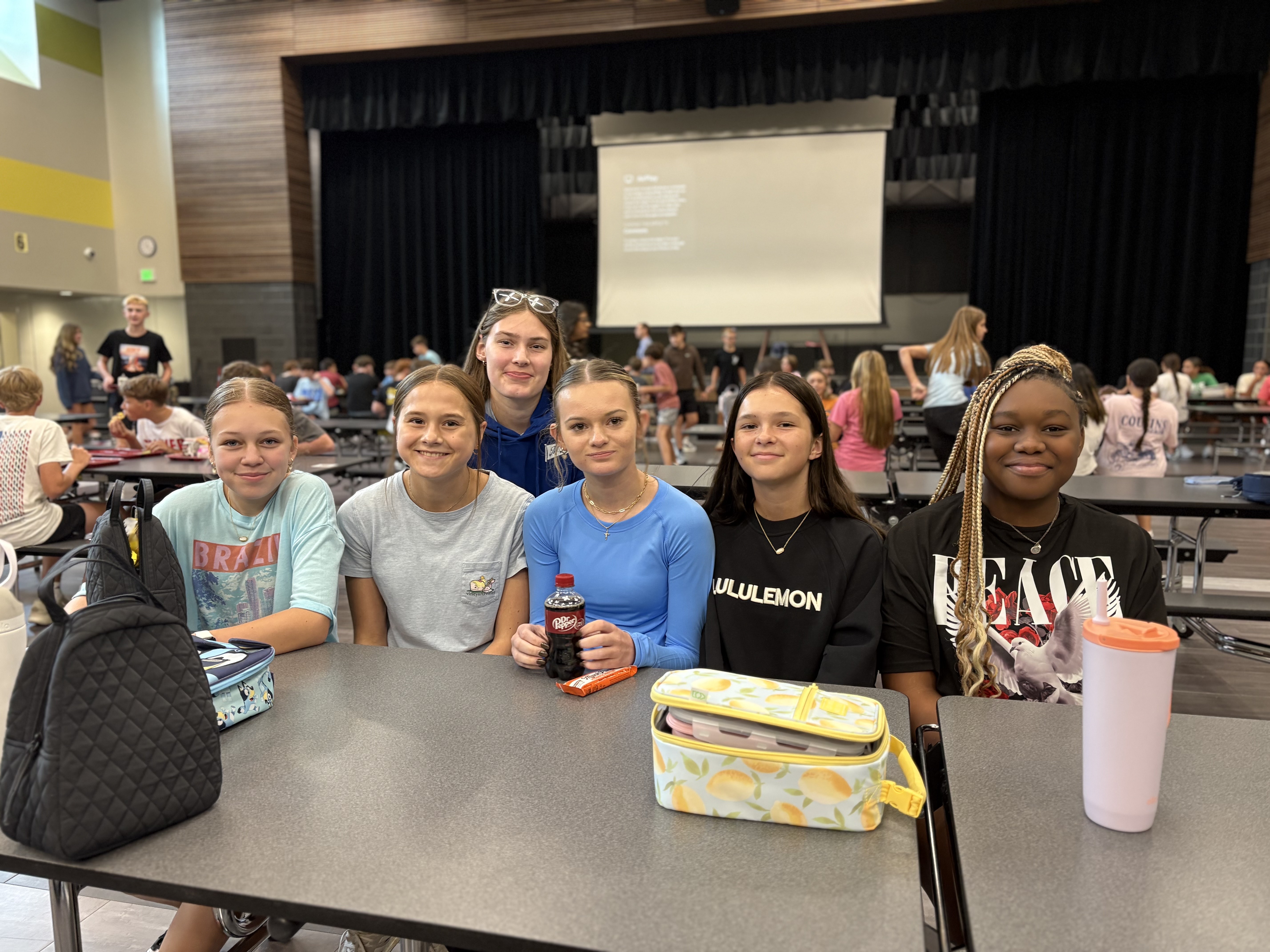 a group of students sitting at the cafeteria table smiling at the camera eating their lunch